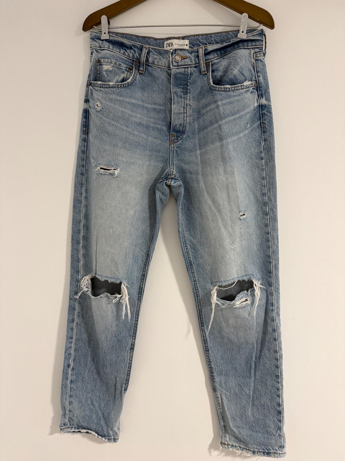 Distressed blue jeans with ripped knees, hanging on a brown hanger against a white background. Visible tag: "Zara".