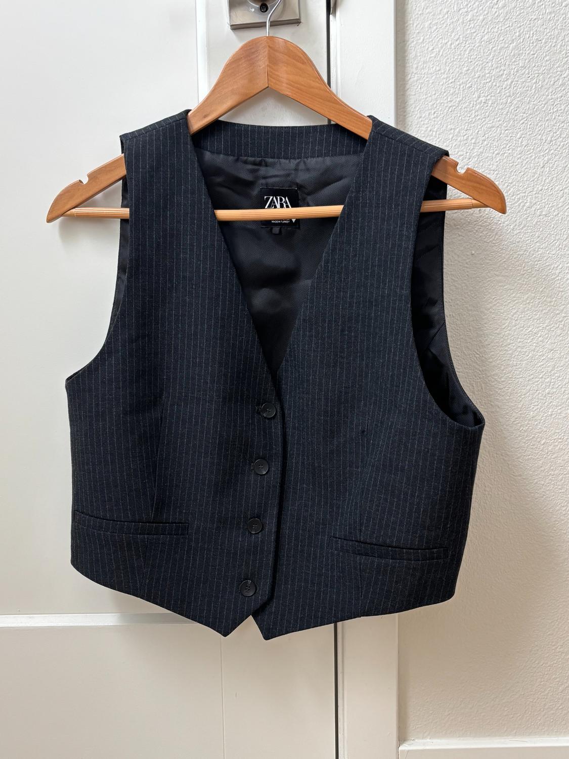 Dark gray dress vest with thin stripes and front buttons, ZARA label, hanging on a wooden hanger over a white door.