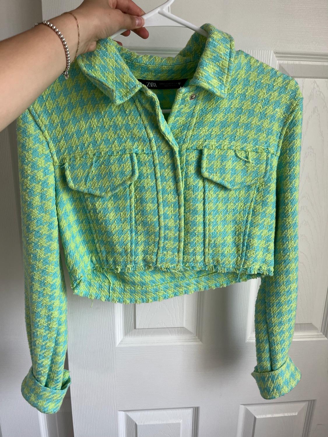 A short jacket with a plaid design in shades of green and yellow is hanging on a white hanger, held by a hand wearing a silver bracelet.