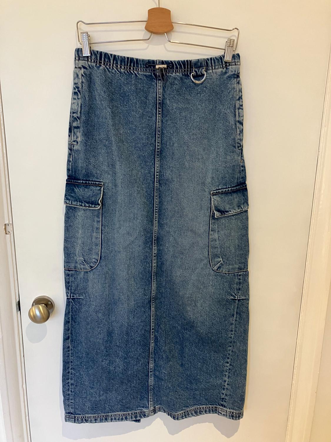 Long blue denim skirt with large pockets, hanging on a wooden hanger over a white door.