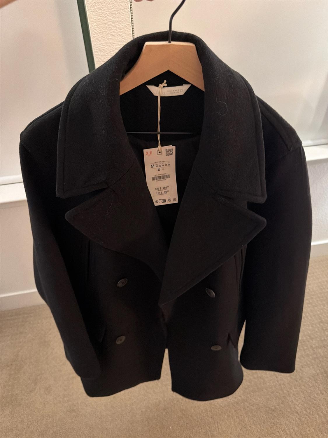 Black double-breasted coat with wide lapels on a wooden hanger. Price tag: US $169.00 UK £99.99. Interior background with a wall and carpet.