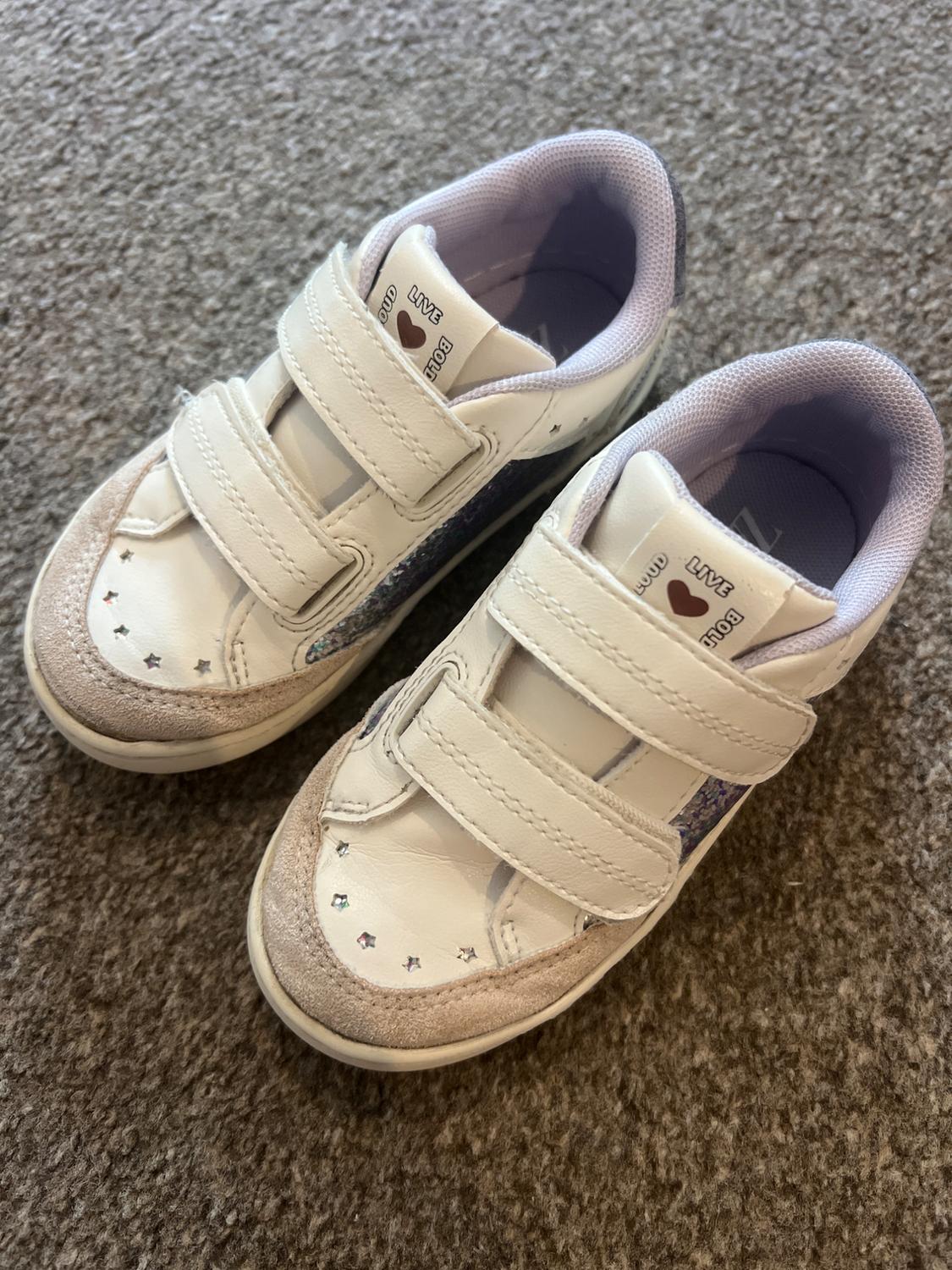 White trainers with hook-and-loop fastenings on a carpet. They feature star details, glitter and the text LIVE LOUD BOLD Z.