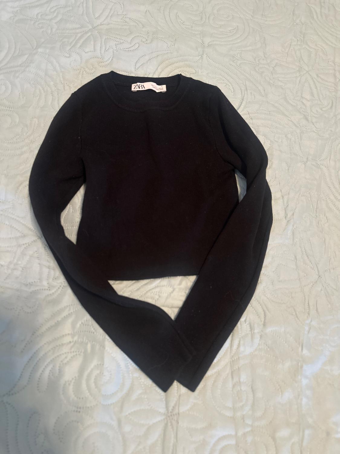 Black long-sleeved jumper with a ZARA label, laid out on a light-coloured bedspread with a curved pattern texture. Relaxed composition.