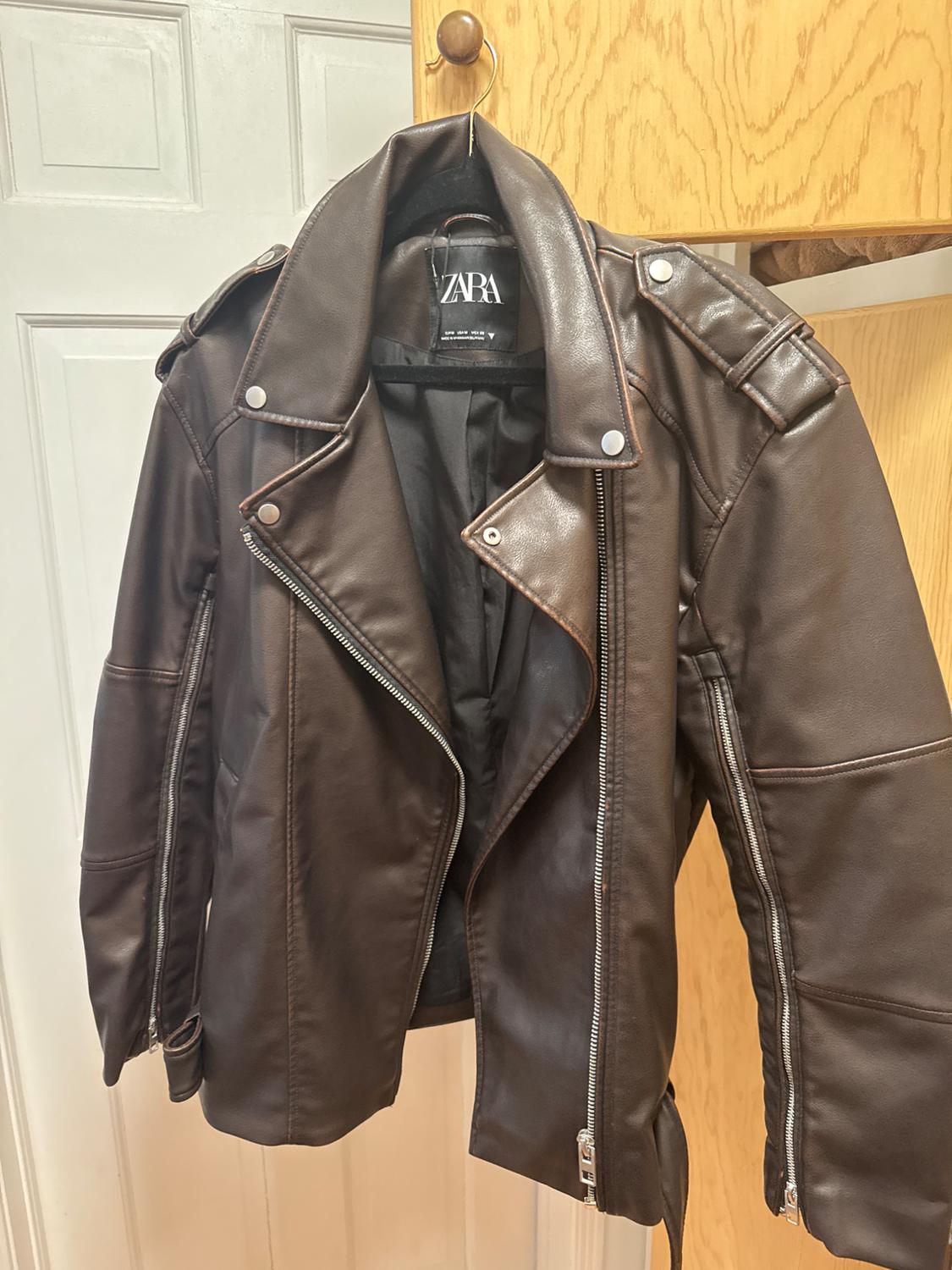 Brown leather jacket hanging on a wooden hook. Silver zip and button details. Visible label with the text "ZARA".