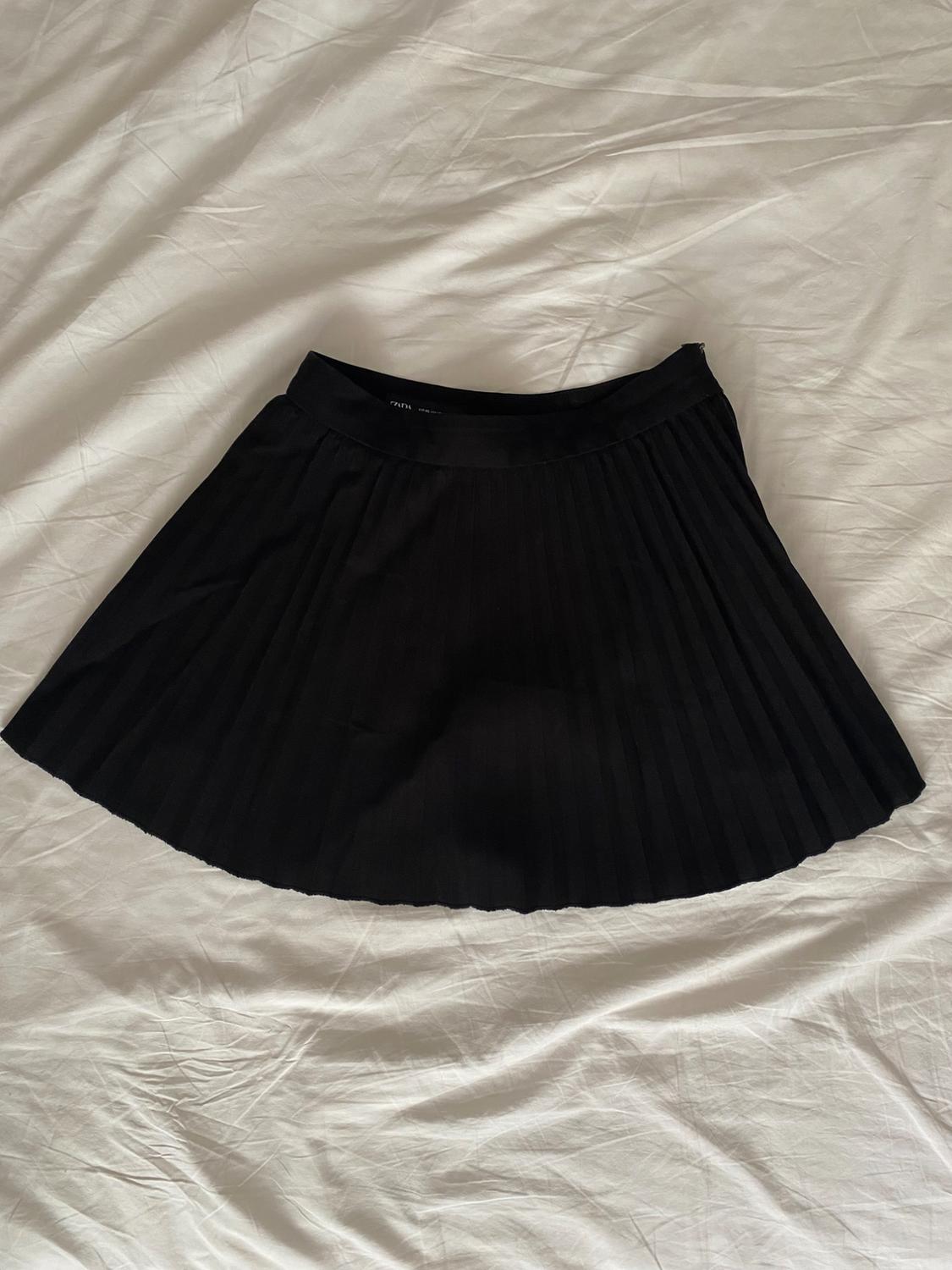 Black pleated mini skirt on a crumpled white sheet. Relaxed overhead shot. ZARA text visible on label.