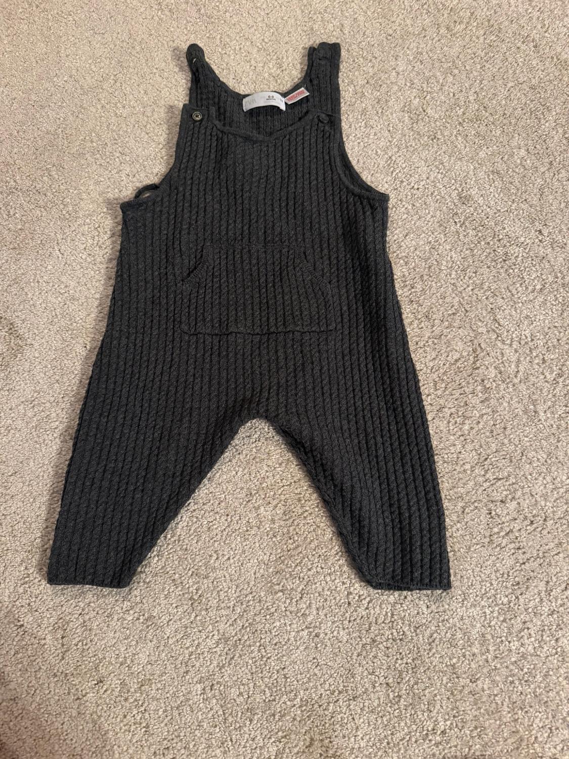 Dark grey knitted baby dungarees with a ribbed texture, buttons on the straps and a central pocket, on a beige rug.