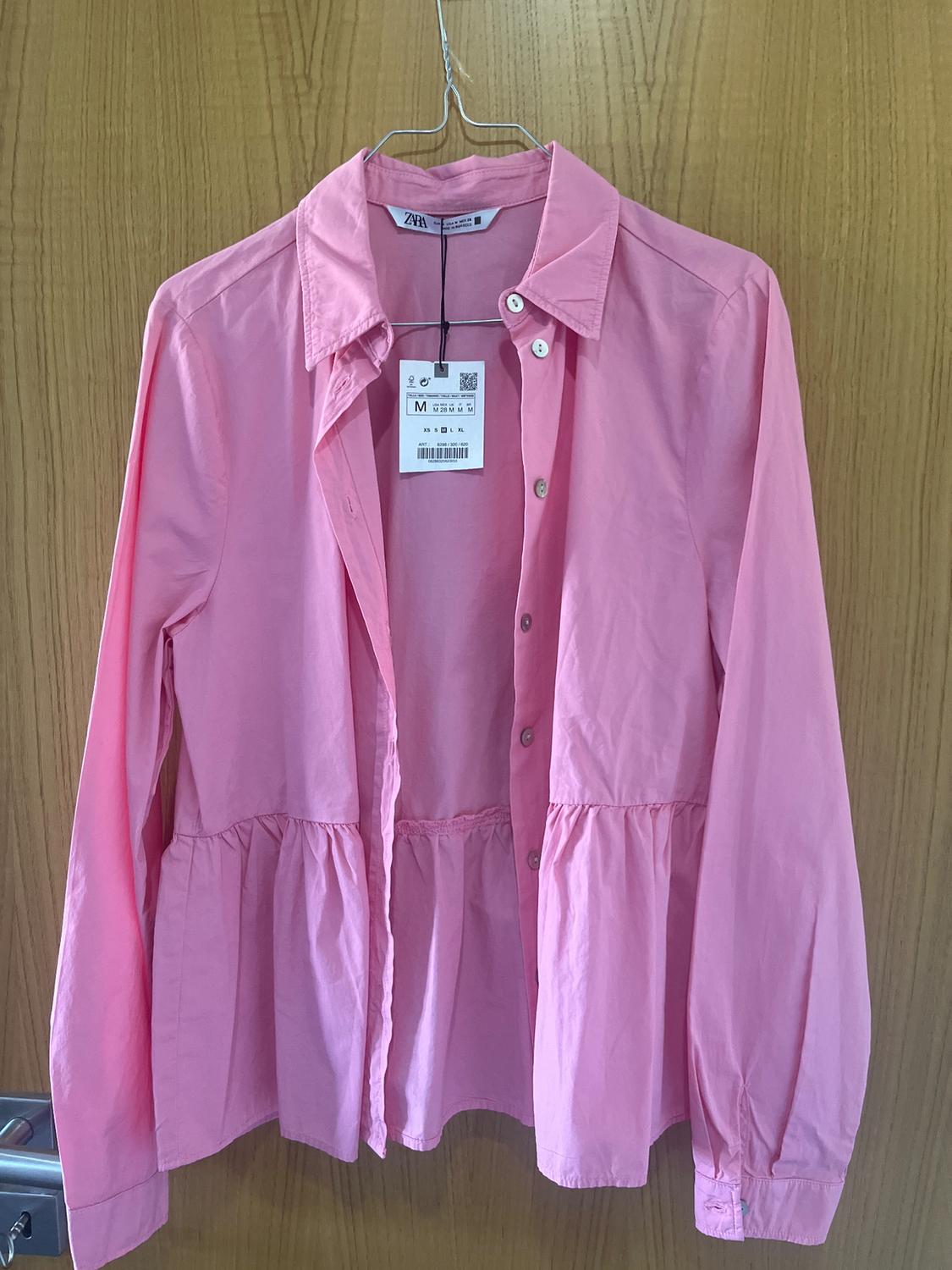 Pink long-sleeved shirt hanging on a metal hanger, with size M label visible, against a light wood background.