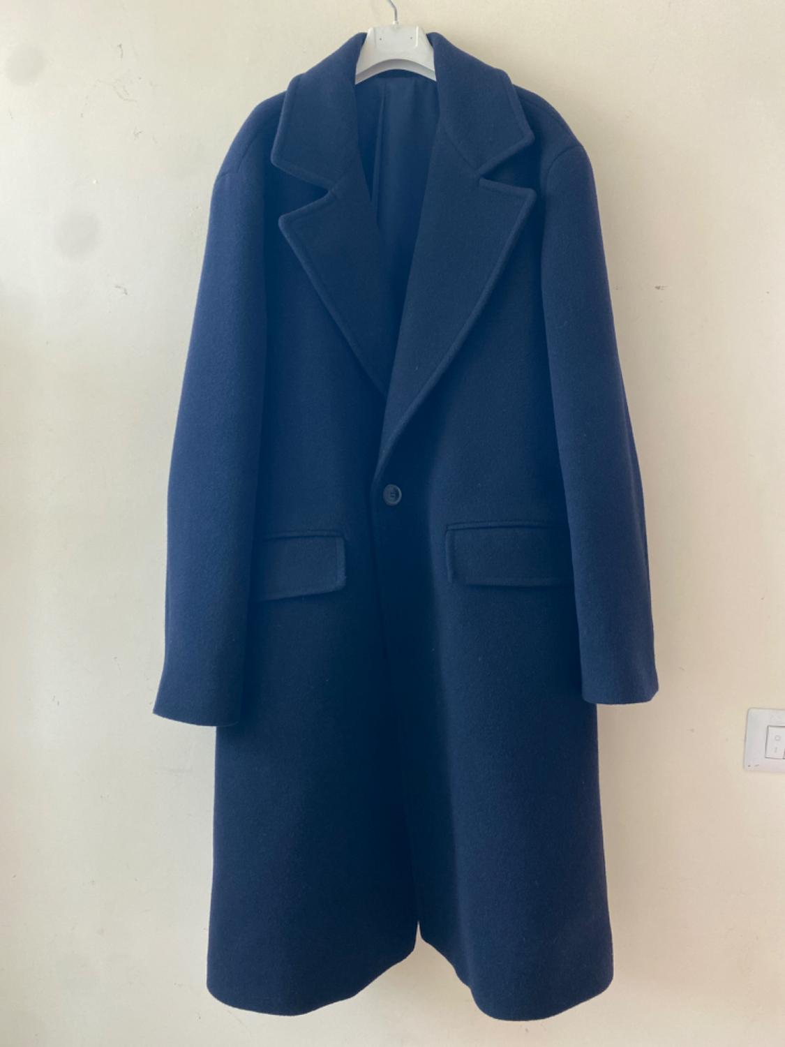 Dark blue coat hanging on a white hanger against a light-coloured wall. Classic design with wide lapels and a visible button.