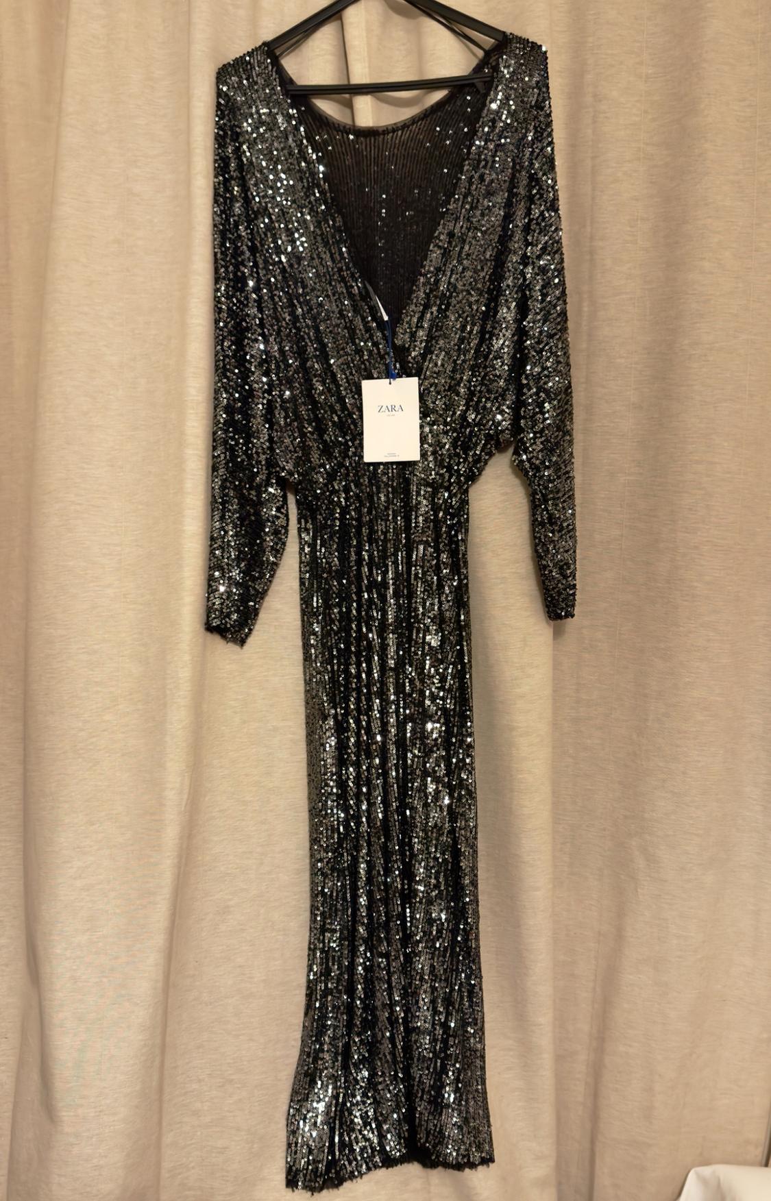 Long black sequinned dress with a V-neckline and long sleeves hanging on a coat hanger in front of a curtain. Label with the text ZARA.
