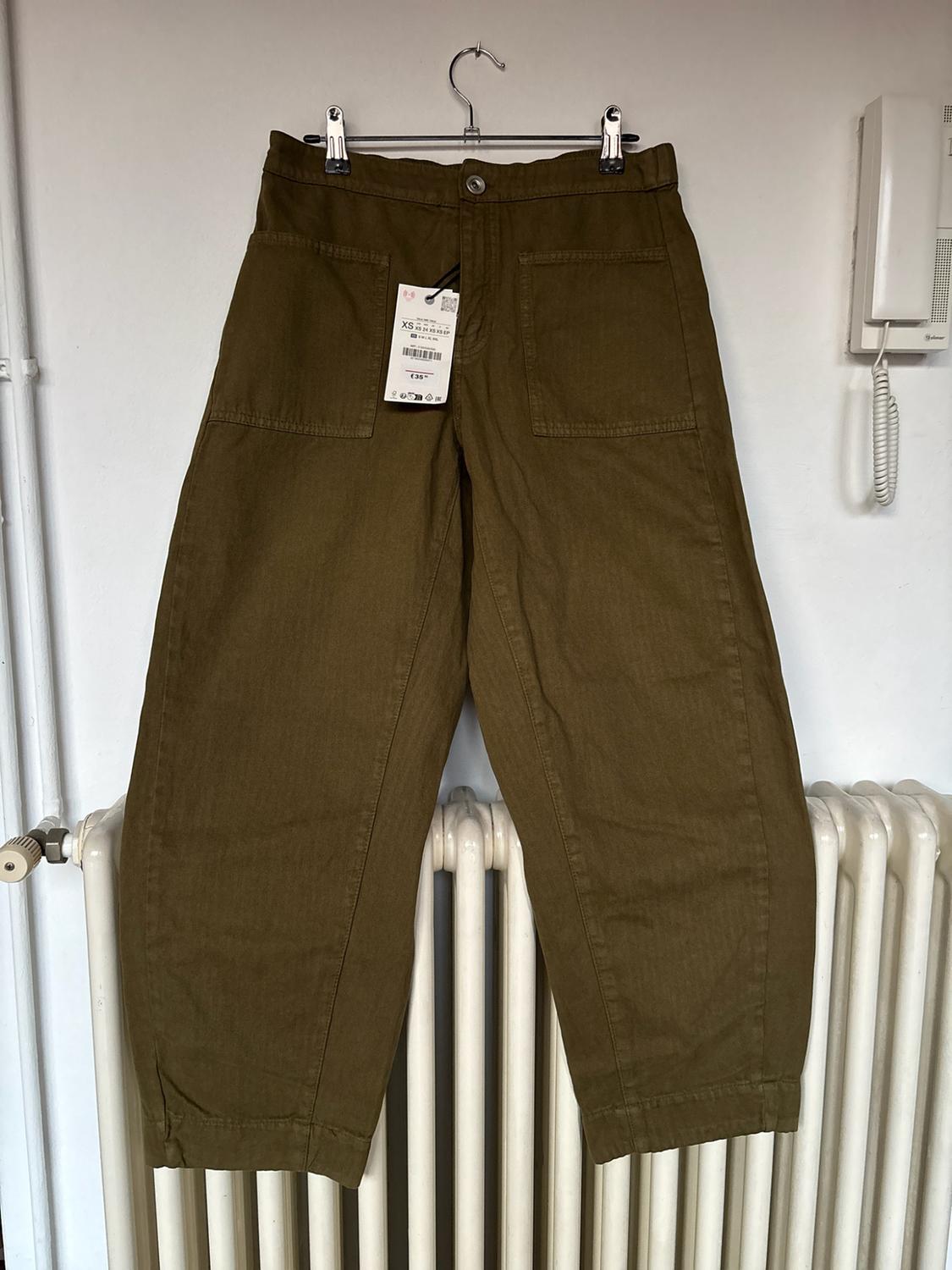 Olive-green trousers with front pockets hanging from a coat hanger above a white radiator. Label with the text XS and €35.