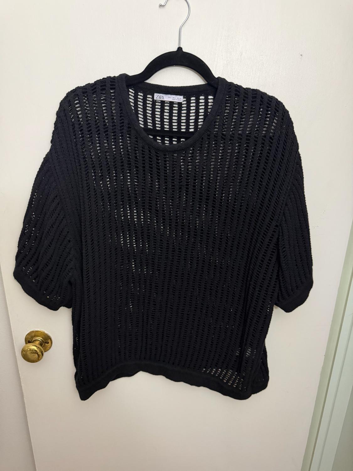 Black open-knit short-sleeve top on a black hanger over a white door with a gold knob. Zara text on the tag.