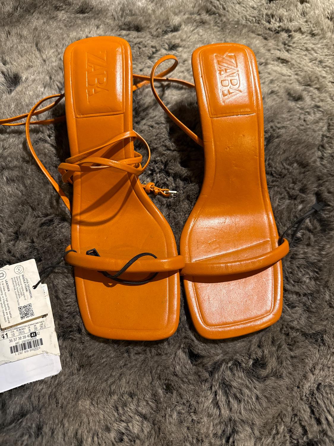 Orange strappy sandals on a gray rug, with a visible logo on the insole. Price tag on the left side.