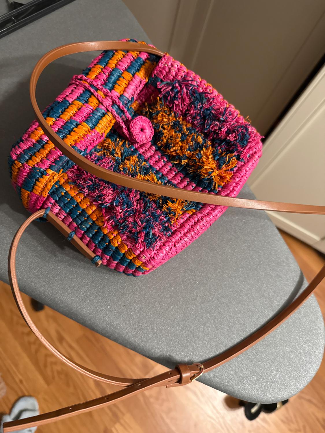 Multicolor woven bag with pink, blue, and orange tones, on a gray table. Adjustable brown strap. Wooden floor visible.
