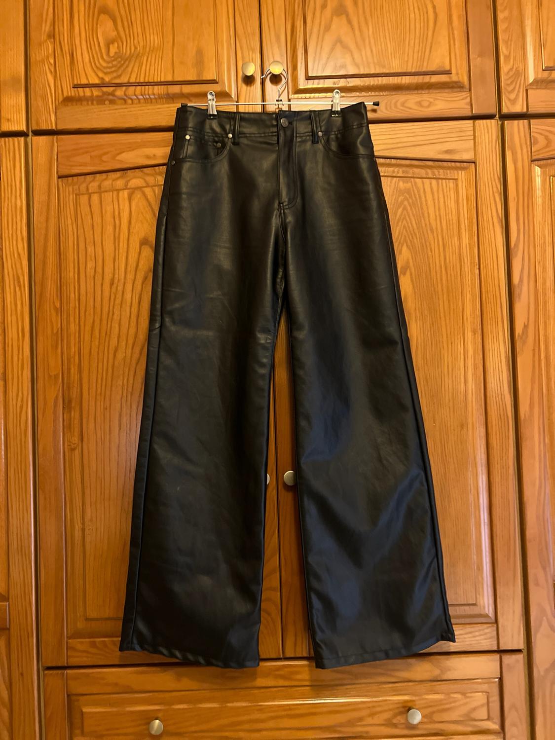 Black wide-leg leather-look trousers hanging on a hanger in front of the doors of a wooden wardrobe.