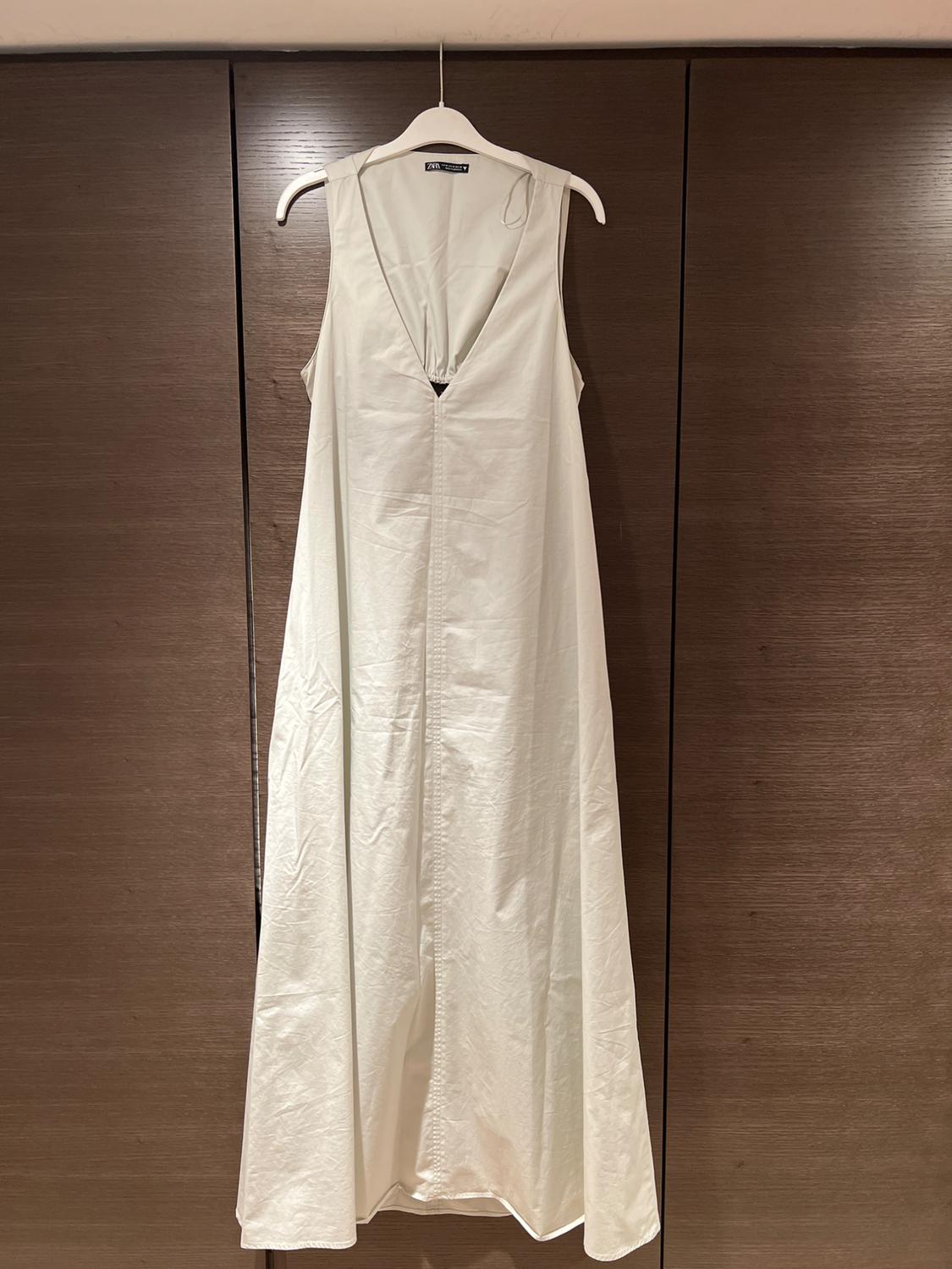 White, sleeveless, V-neck maxi dress, hanging on a white hanger in front of dark brown wooden wardrobe doors.