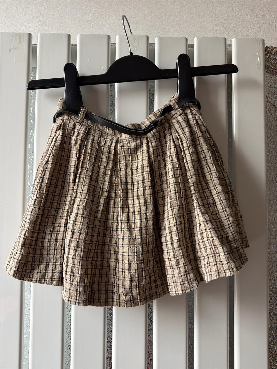 Short pleated beige and black check skirt with a black belt, hanging on a hanger in front of a white radiator.