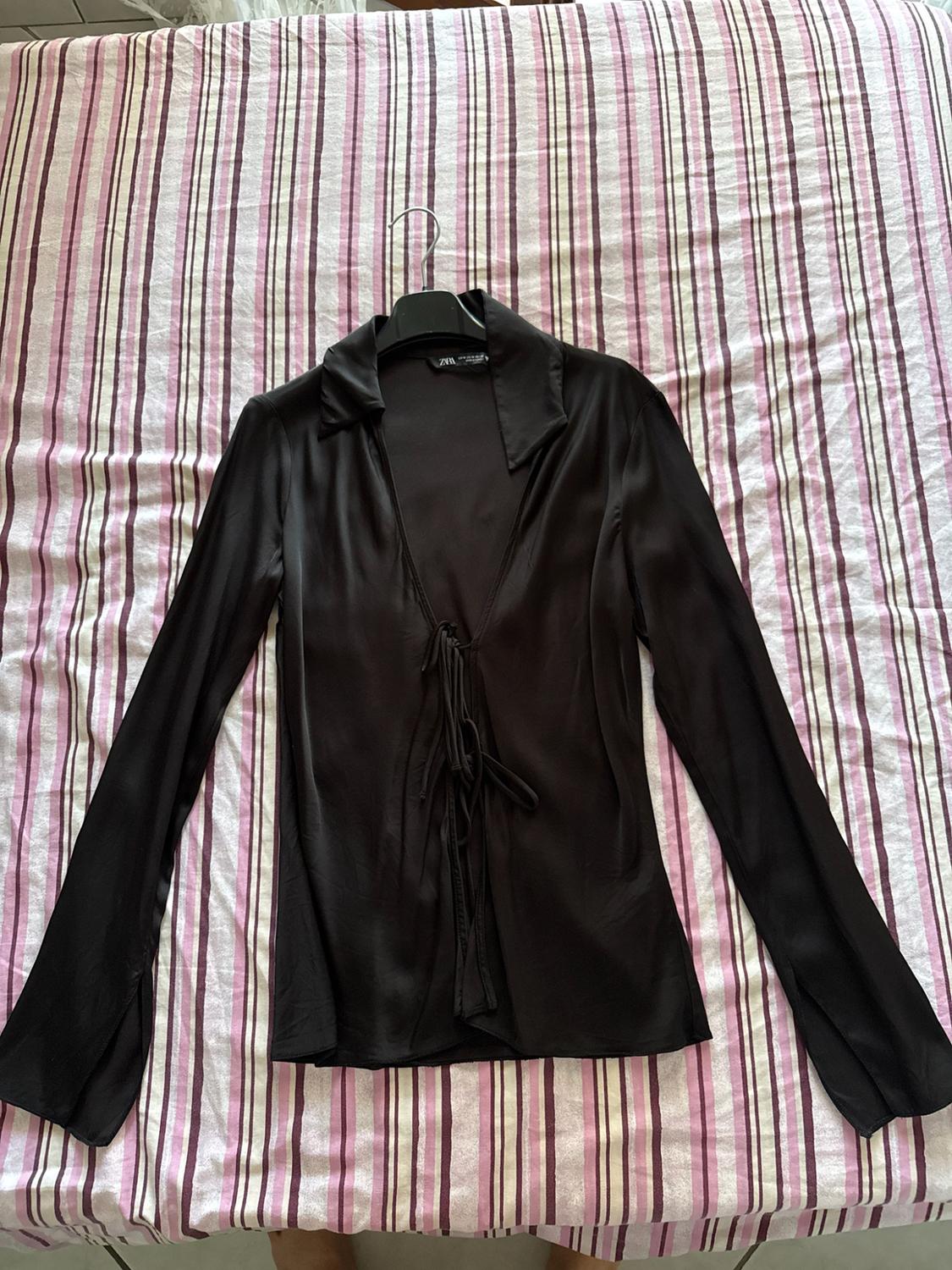 Black long-sleeved shirt with a collar and front tie, on a hanger on a bed with striped sheets. Visible text: ZARA.