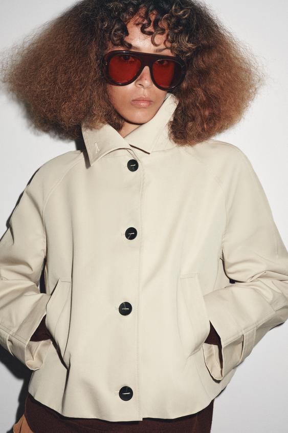 "Středně dlouhý béžový krátký trenčkot se šálovým límcem, černými předními knoflíky a předními kapsami."