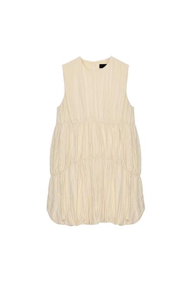 Front view of a short, sleeveless, cream dress with a round neck, gathers, and a balloon hem. Concealed side pockets.