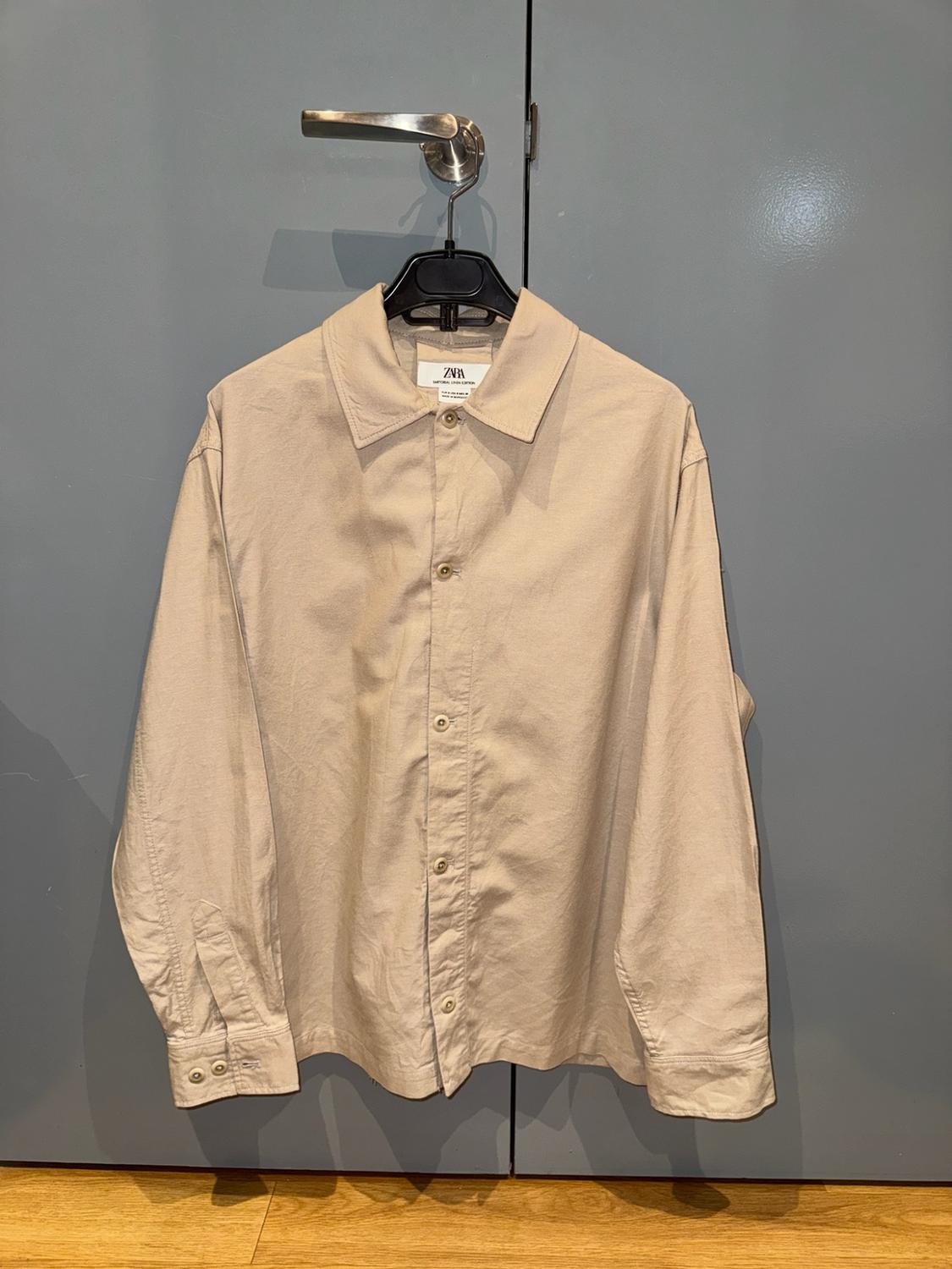 Beige long-sleeved shirt with buttons and ZARA label, hanging on a hanger from the metal handle of a grey wardrobe.
