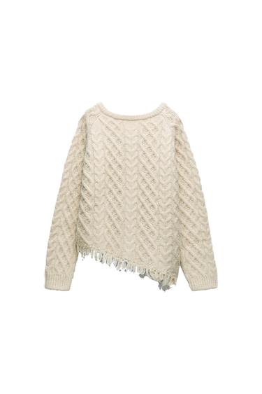 Cream chunky knit sweater with cable-knit design and fringe on the bottom hem, on a white background.