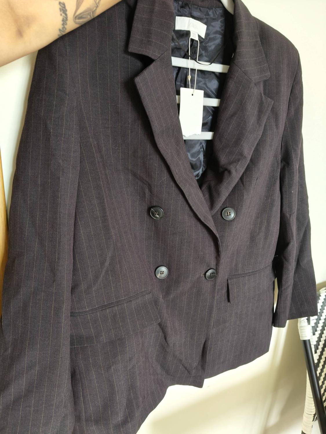 Tattooed arm with “Morriña” holds a dark, double-breasted pinstripe blazer on a hanger with a label, against a white background.