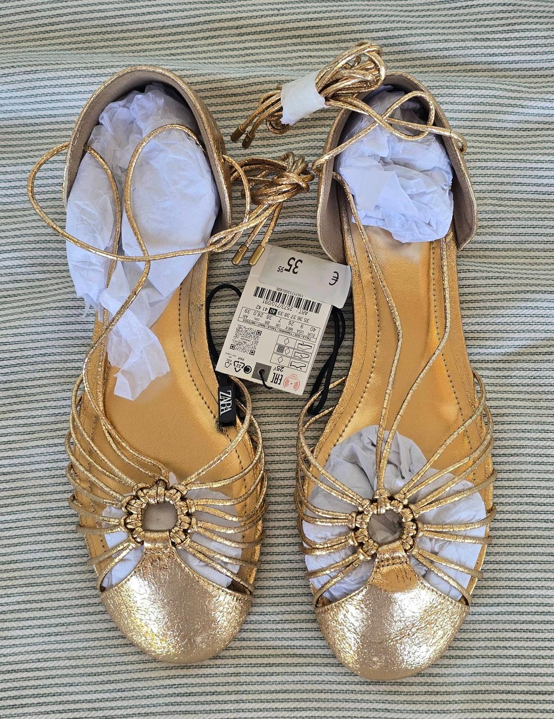 Gold strappy flat sandals on striped fabric. They have paper inside and a tag with the text ZARA and € 35.95.