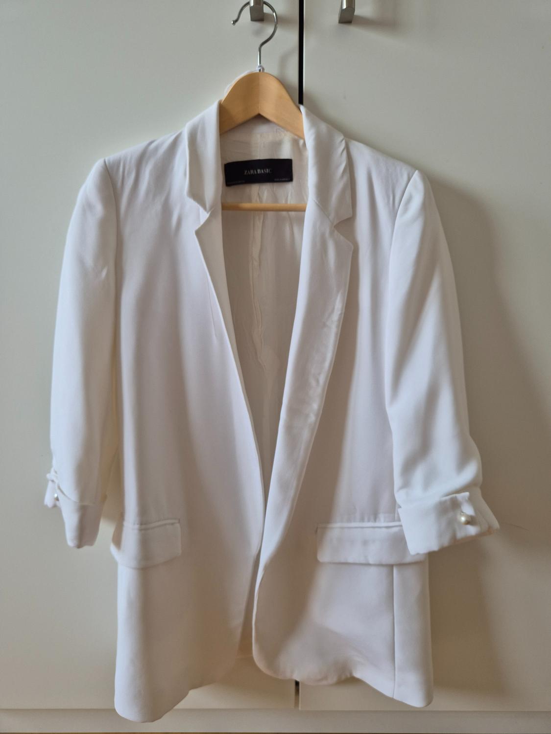 White blazer hanging on a wooden hanger, with a visible label that reads "ZARA BASIC", against a white wardrobe background.