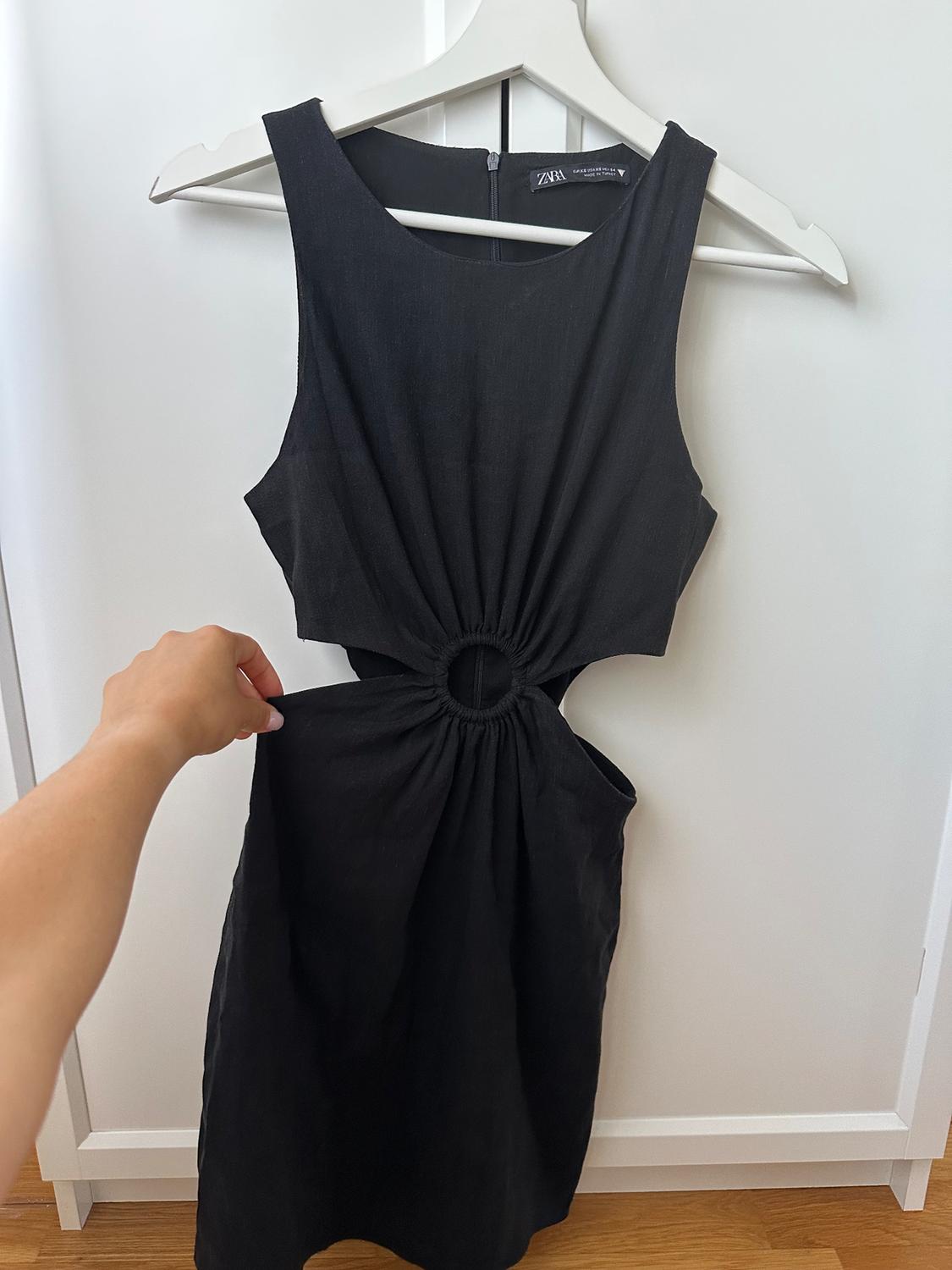 A sleeveless black dress with a ring detail at the waist is hanging on a white hanger. A hand is holding the dress.
