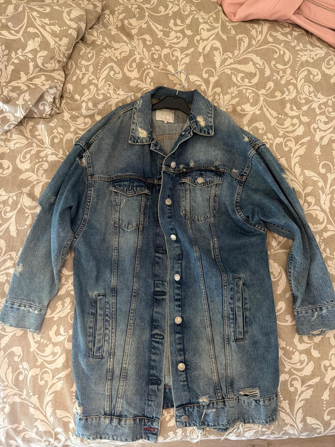 Blue denim jacket with distressed and ripped details, laid out on a bed with a floral-patterned bedspread.