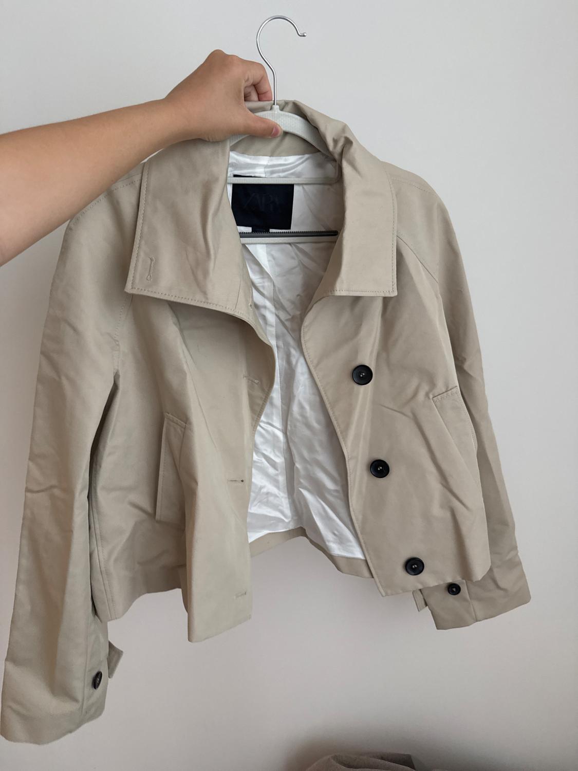 Hand holding a short beige jacket on a hanger against a white background. Open, it shows black buttons, a white lining, and a ZARA label.