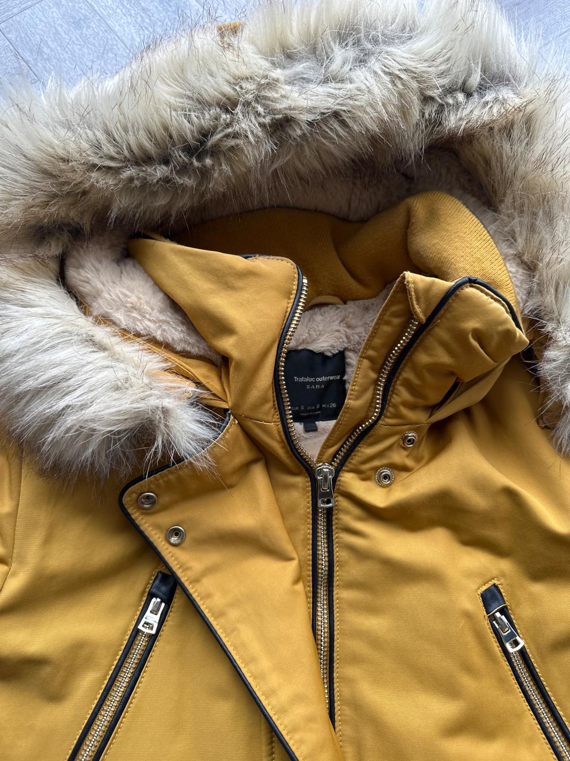 Mustard coat with faux fur hood and black zips. Visible label: "trafaluc outerwear ZARA". Light grey background.