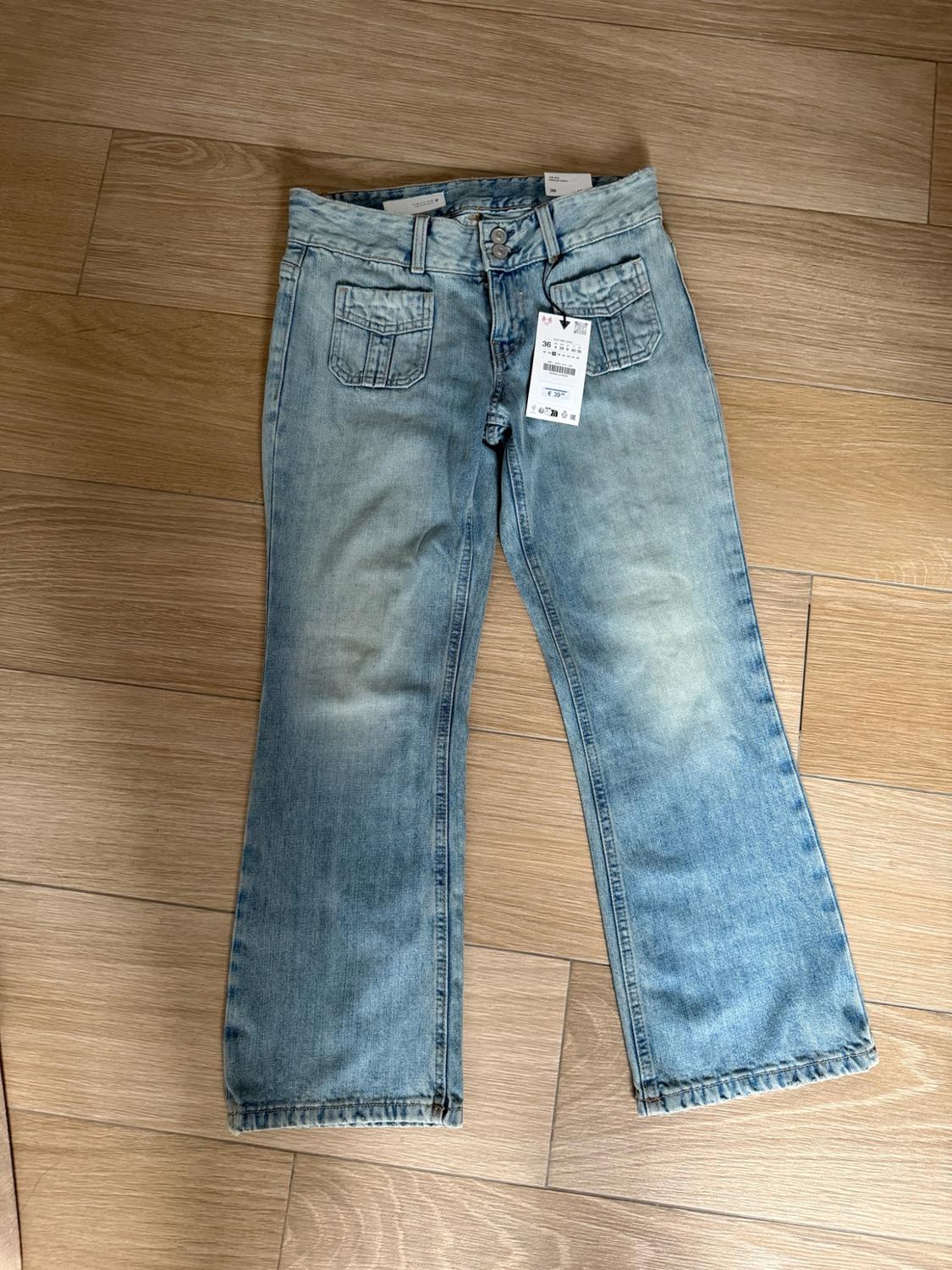 Light blue denim jeans with front flap pockets and a label, laid out on a wooden floor.