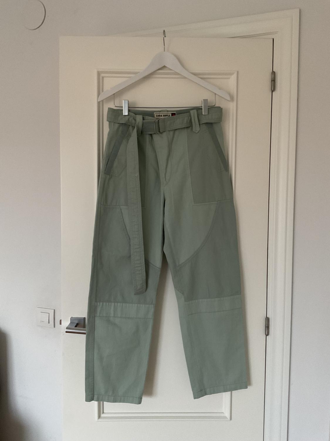 Green trousers hanging on a white hanger over a white door. The label reads "ZARA SRPLS".