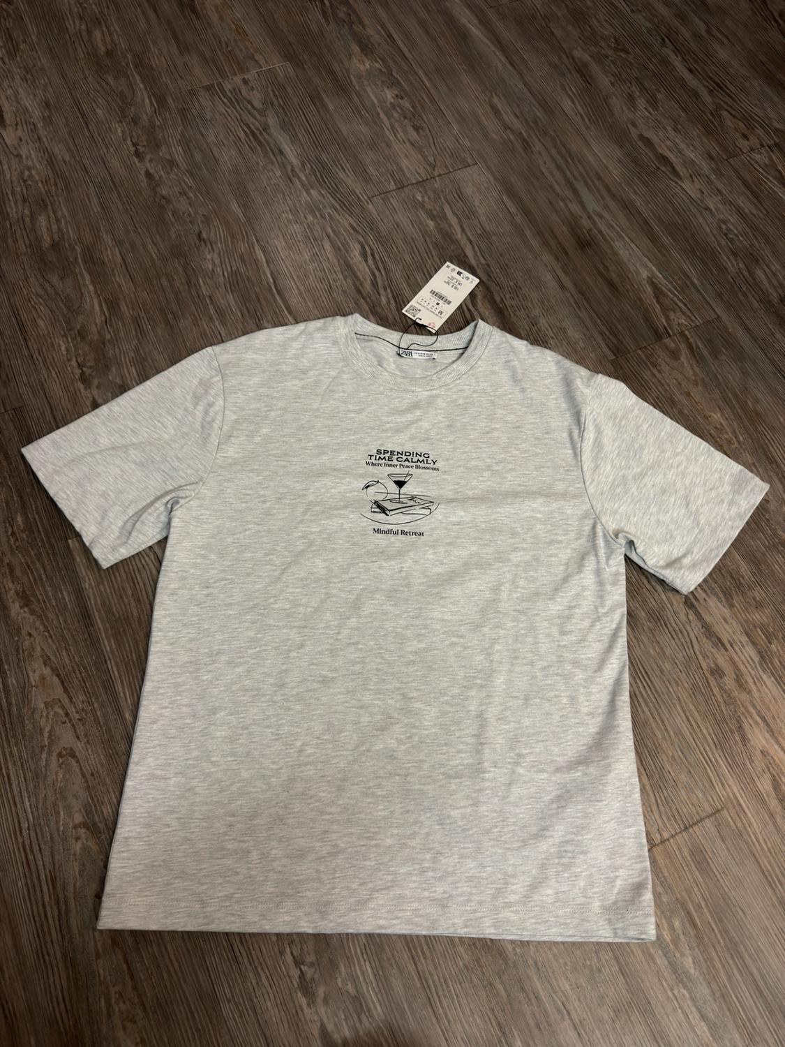 Grey T-shirt on wood with a graphic of a cup and book. Text: SPENDING TIME CALMLY Where Inner Peace Blossoms Mindful Retreat.