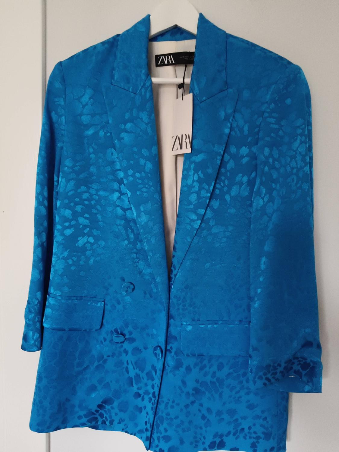 Blue jacket with an abstract print, hanging on a white hanger. Visible label with the text "ZARA".