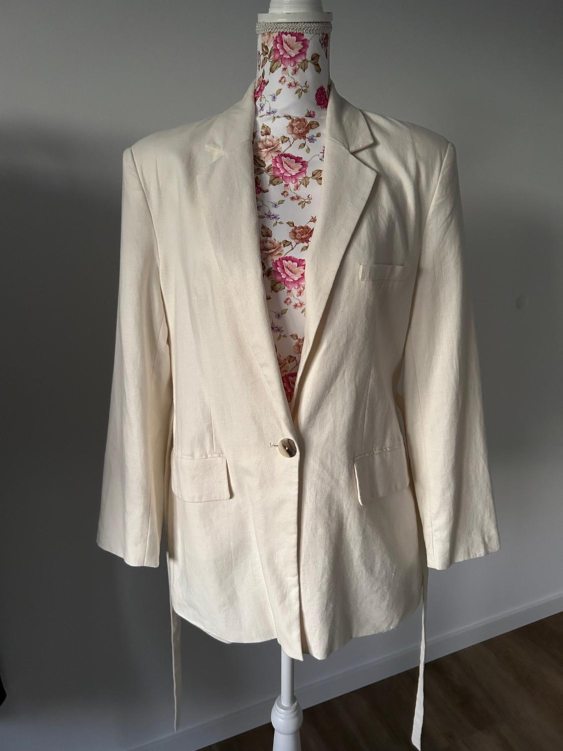 Mannequin with a single-button beige blazer, floral background in pink and brown tones, indoor setting with soft lighting.