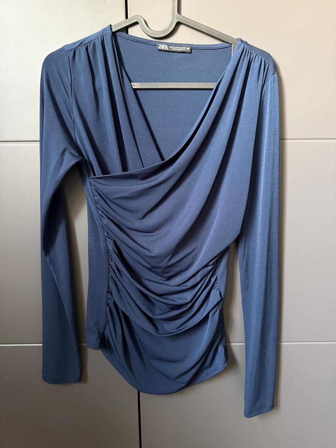 Blue long-sleeved blouse with front draping, hanging on a black hanger against a light background. Label visible.