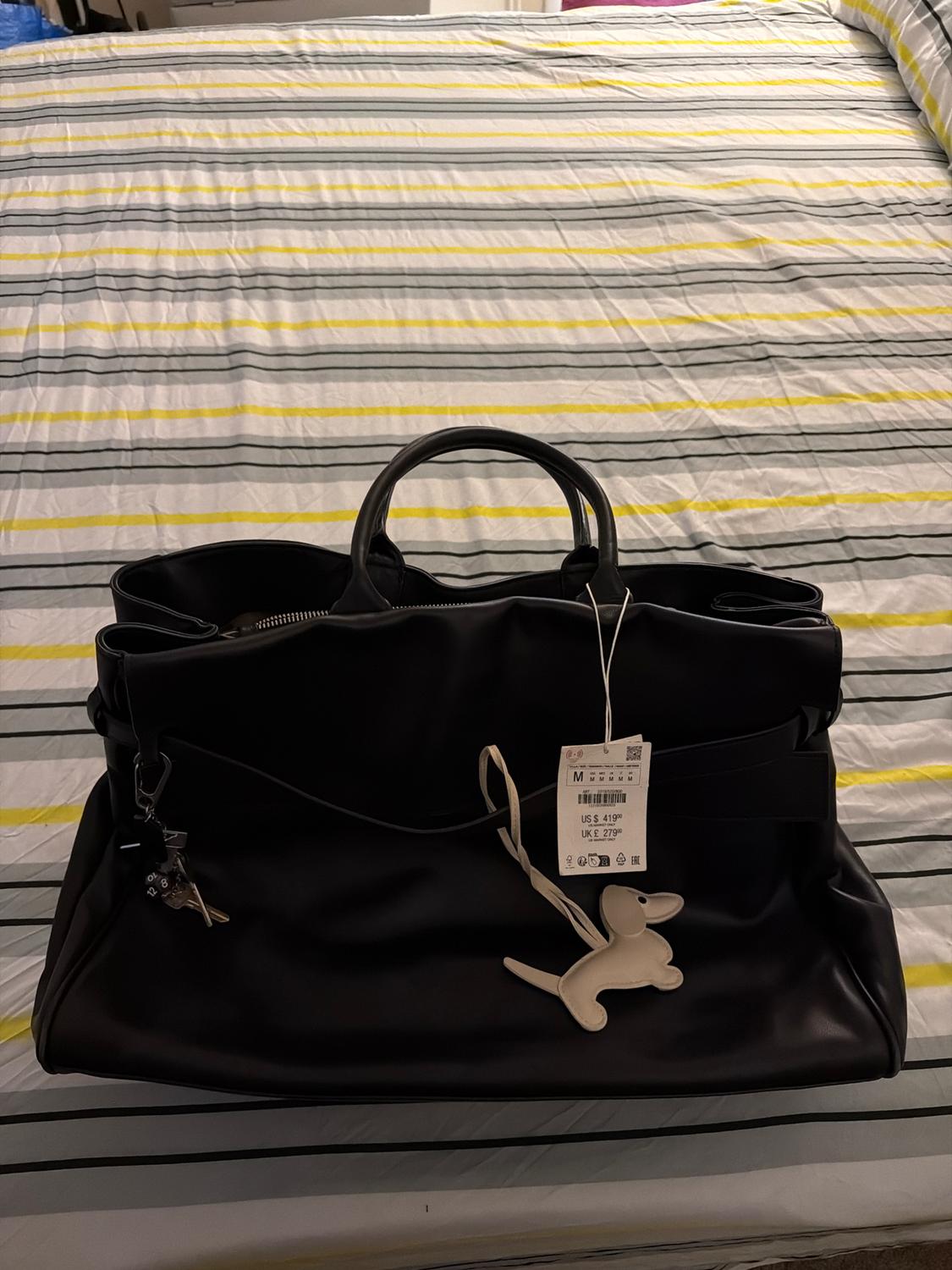 Large black handbag on a bed with a grey and yellow striped duvet. Hanging tag and white dog keyring.
