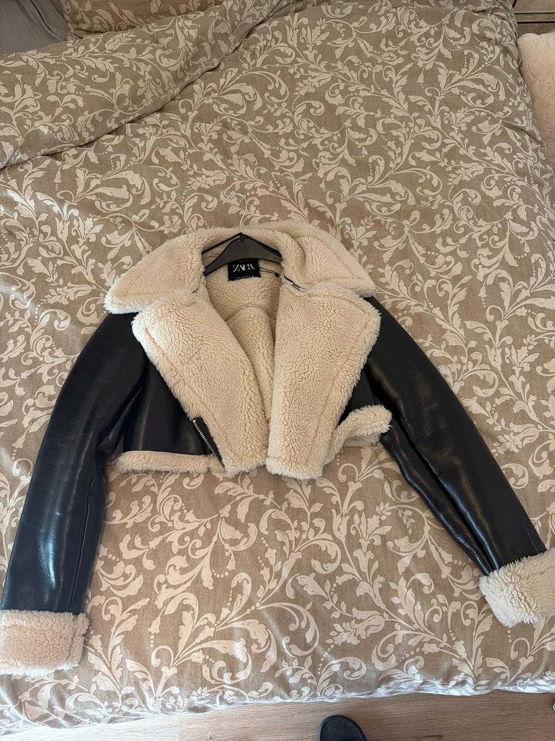 Short black jacket with light-coloured fur lining on a bed with a beige floral-patterned bedspread. ZARA label visible.