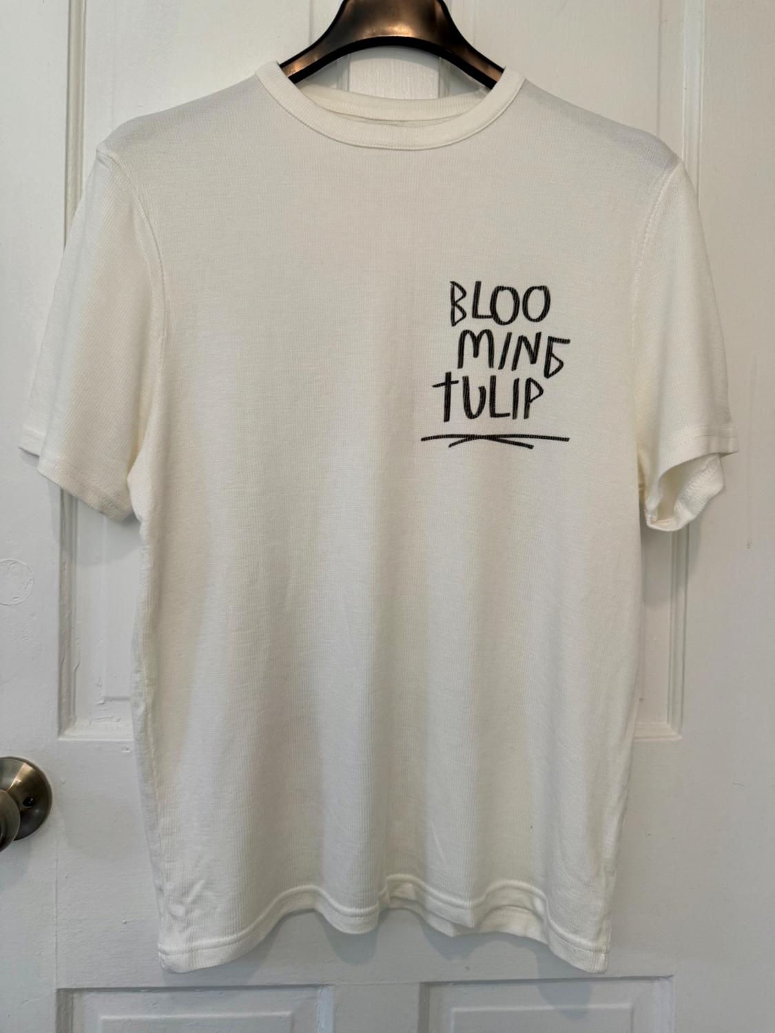 White T-shirt hanging on a black hanger, with "BLOOMING TULIP" text in black letters on the chest, over a white door.