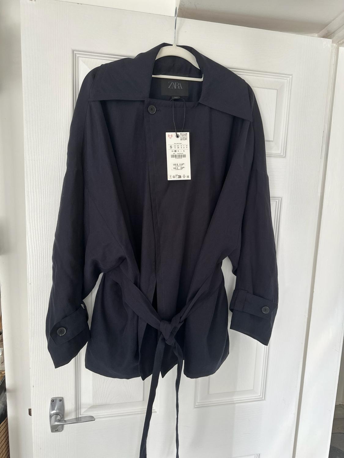 Dark jacket with a belted waist hanging on a white door. You can see zara on the inner label and S on the hang tag.