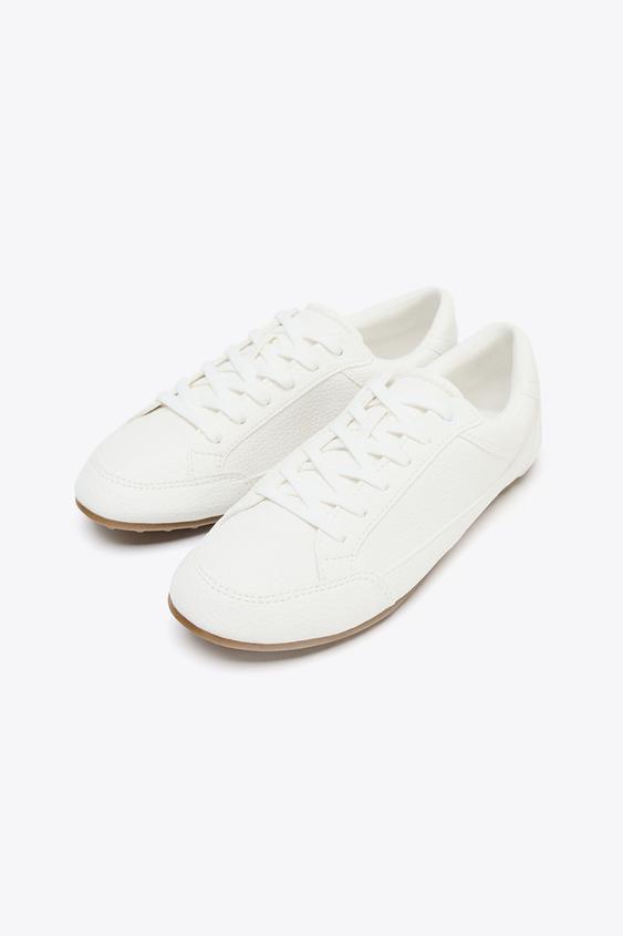 Bílé tenisky s nízkým profilem, tkaničkami a světle hnědou podrážkou.