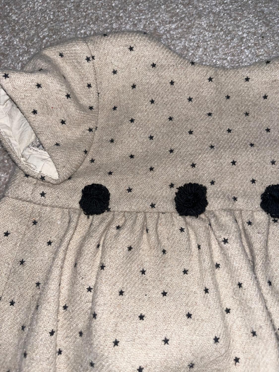 Beige dress with a small black star print and three black pom-poms on the top, on a grey background.
