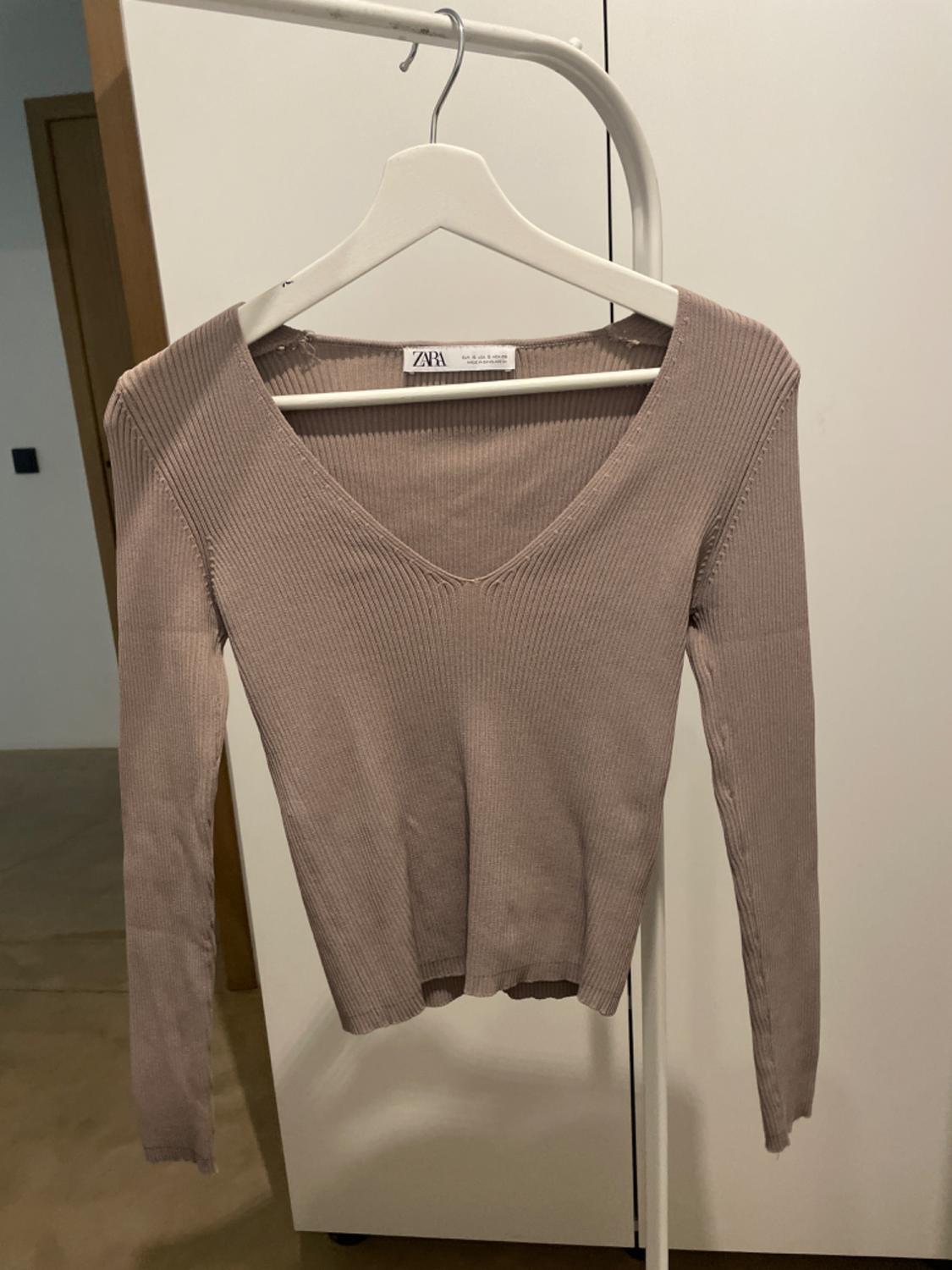 Beige knitted V-neck jumper hanging on a white hanger. Visible label with the text "ZARA". White wardrobe background.