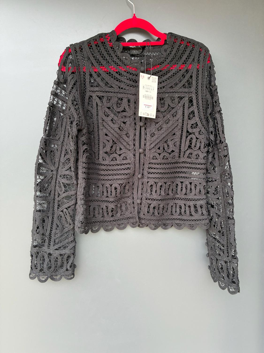 Dark grey lace jacket with long sleeves and buttons, on a pink hanger against a grey background. Text: ZARA S €49.95.