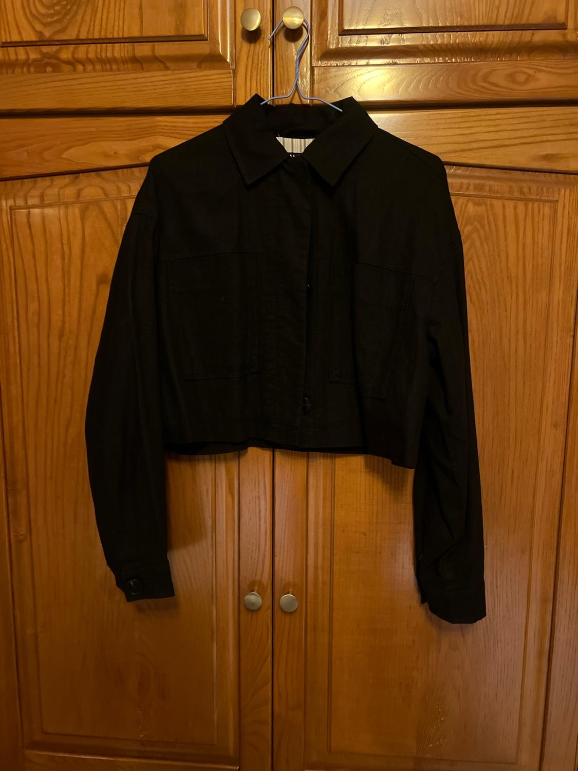 Short black jacket with a collar and front pockets, hanging on a hanger in front of wooden wardrobe doors.