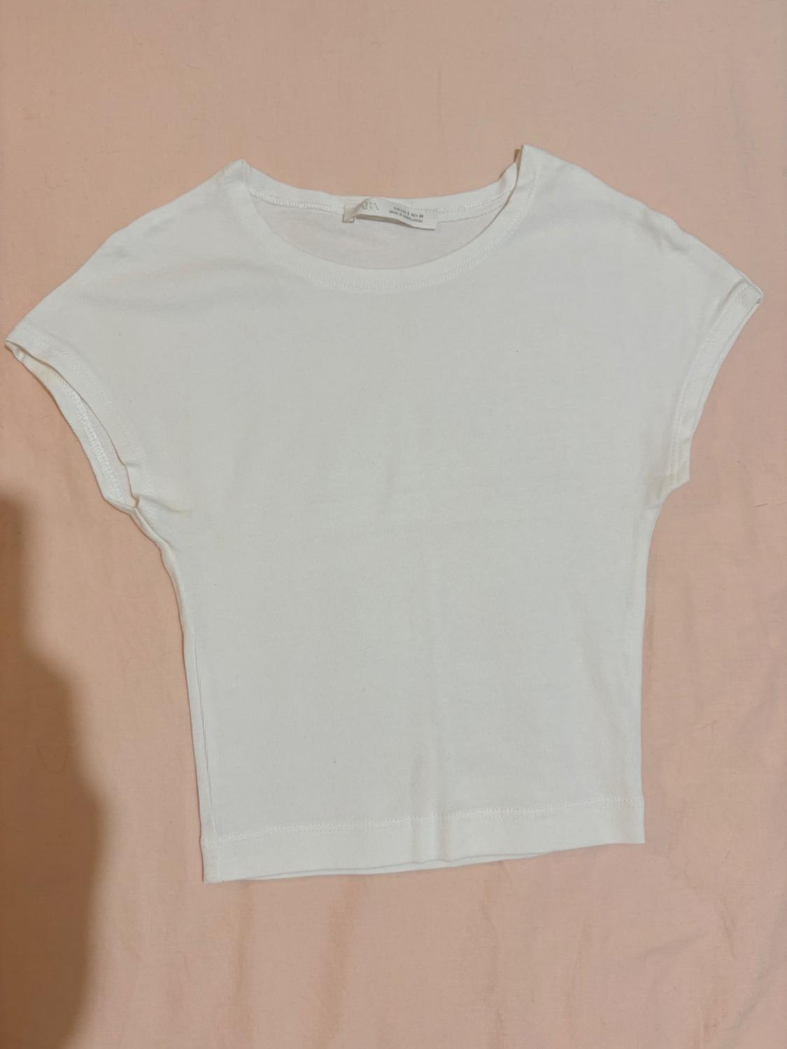 Basic white short-sleeved crew-neck T-shirt, laid flat on a light pink fabric background.