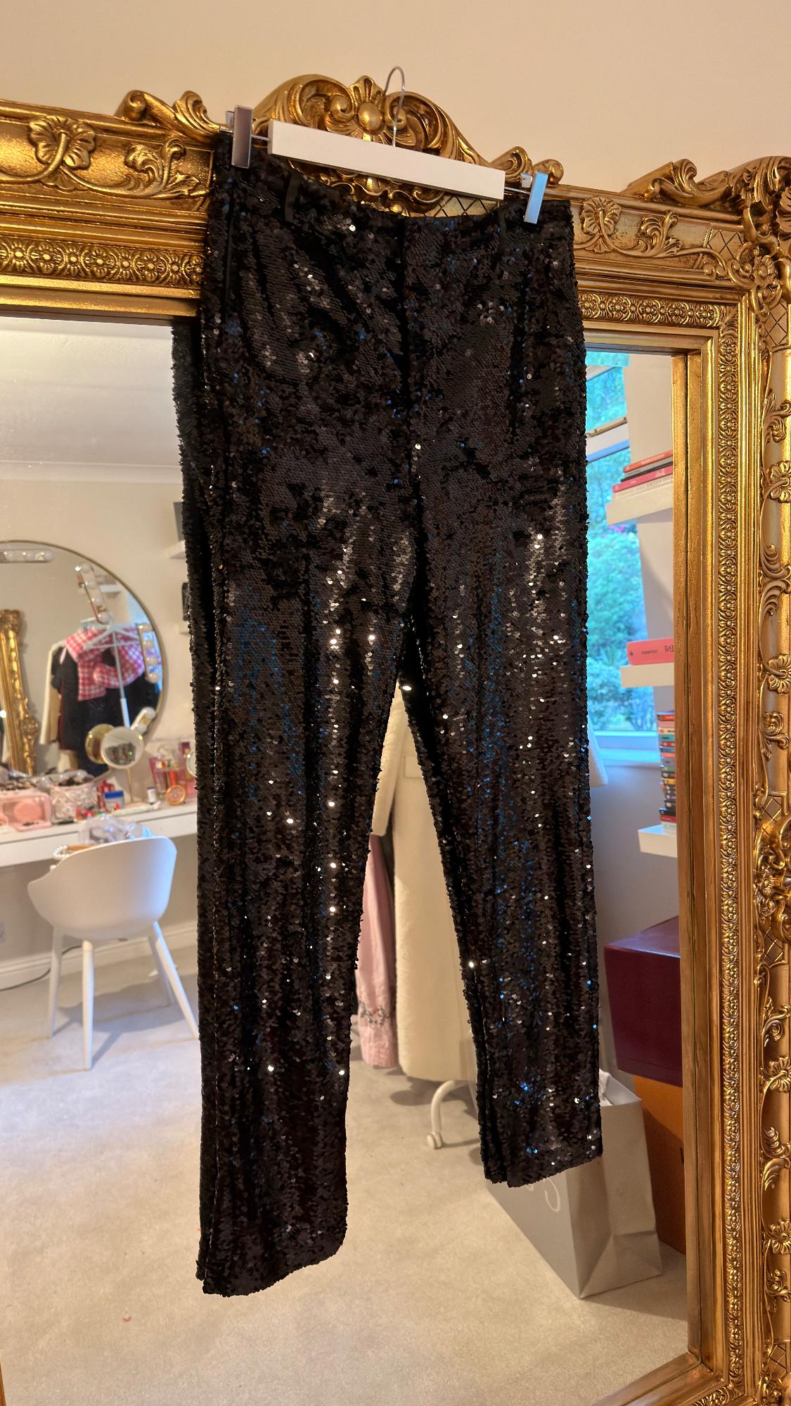 Black sequinned trousers hanging on an ornate gold frame. Mirrored background, white chair and shelving unit.