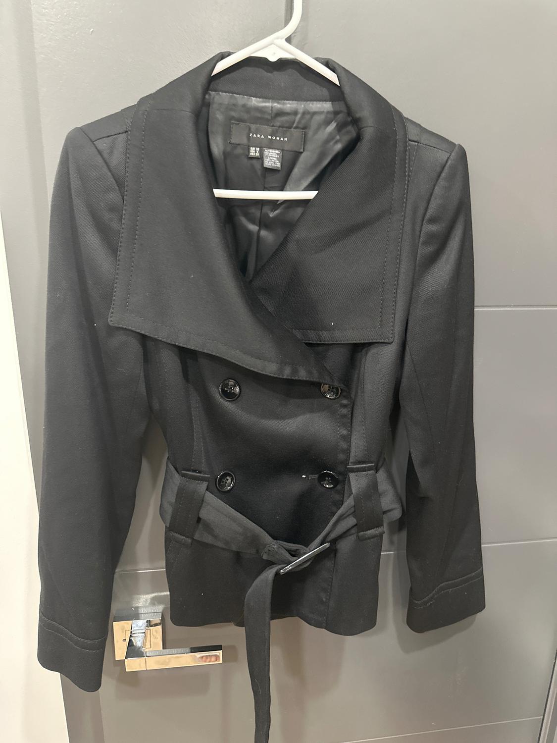 Women's black double-breasted jacket with a belt, hanging on a hanger over a gray door. Visible tag: "ZARA WOMAN".